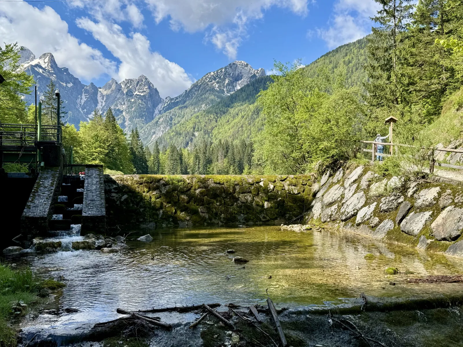 Klares Gewässer mit kleiner Staumauer bei den Laghi di Fusine, Bergpanorama im Hintergrund.