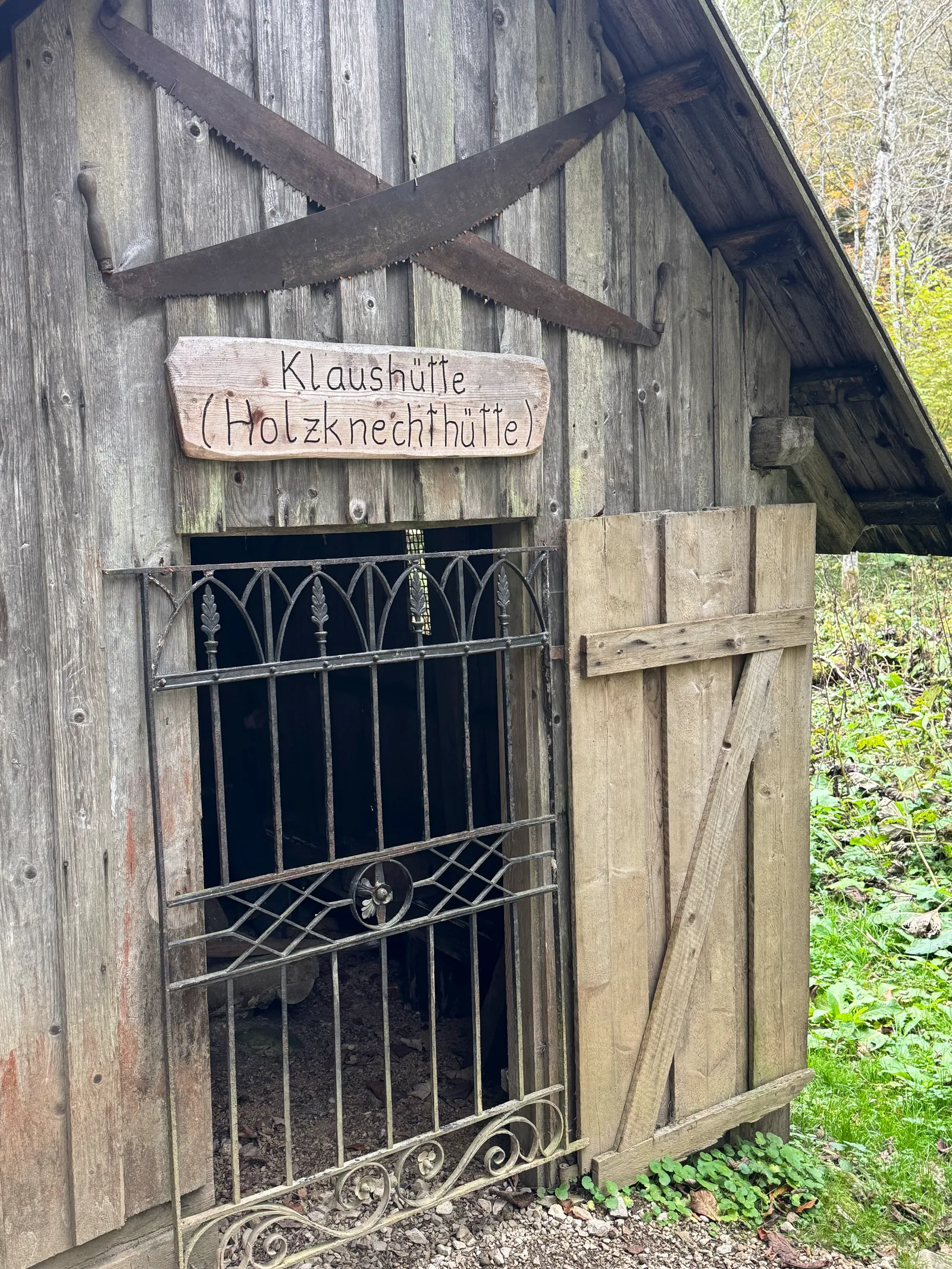 Eingang der Klaushütte (Holzknechthütte) mit altem Holzrahmen, gekreuzten Sägen und schmiedeeisernem Gitter.