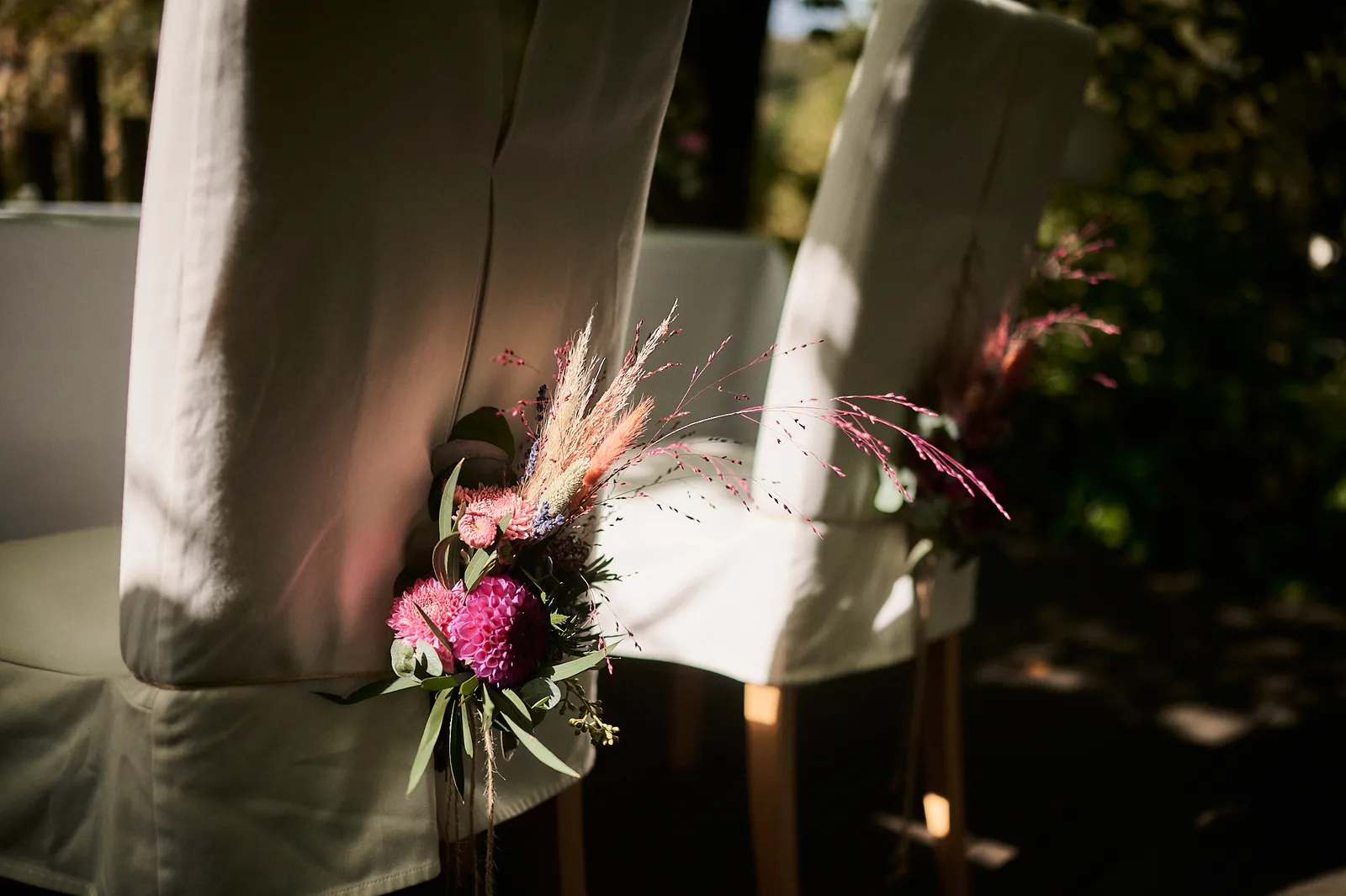 Mit pinken Blumen und Pampasgras geschmückte Stühle bei einer Hochzeitsfeier im Freien.