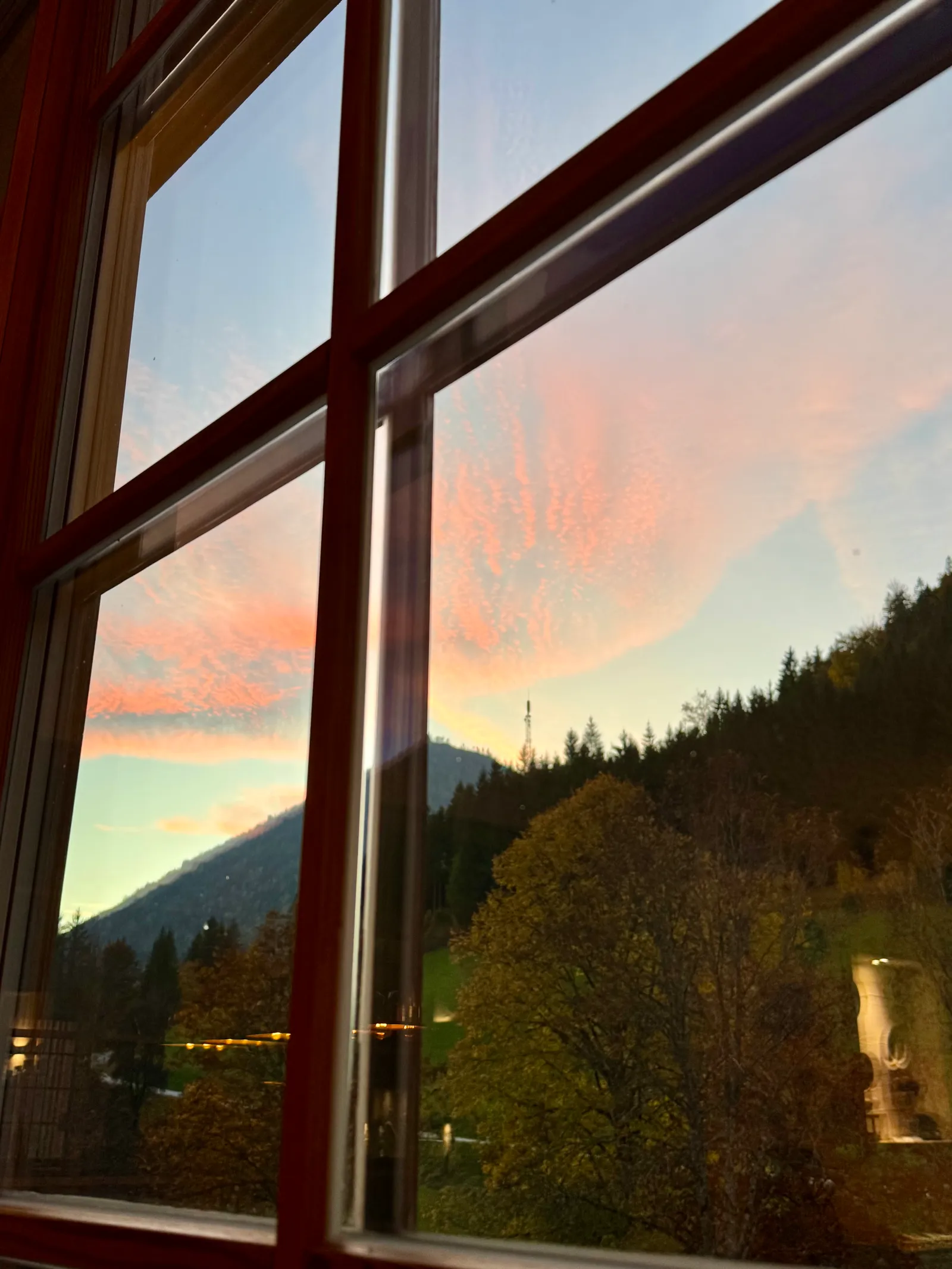 Abendhimmel mit rosa Wolken über Bergen und Bäumen, durch ein Hotelfenster gesehen.