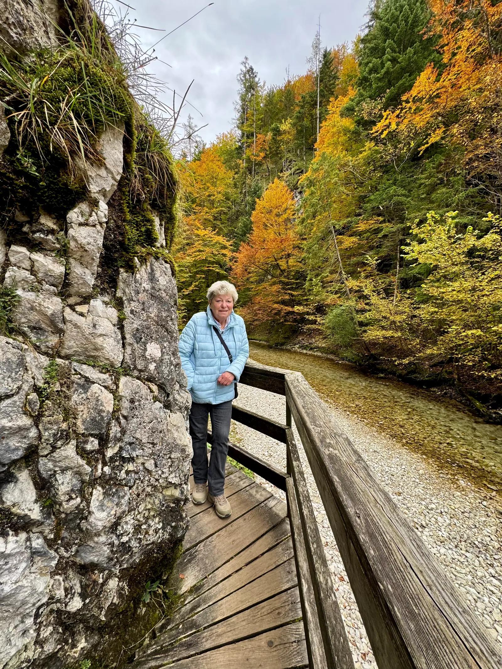 Eine Frau in hellblauer Jacke steht auf einem Holzsteg neben Fels und herbstlichem Bach im Mendlingtal.