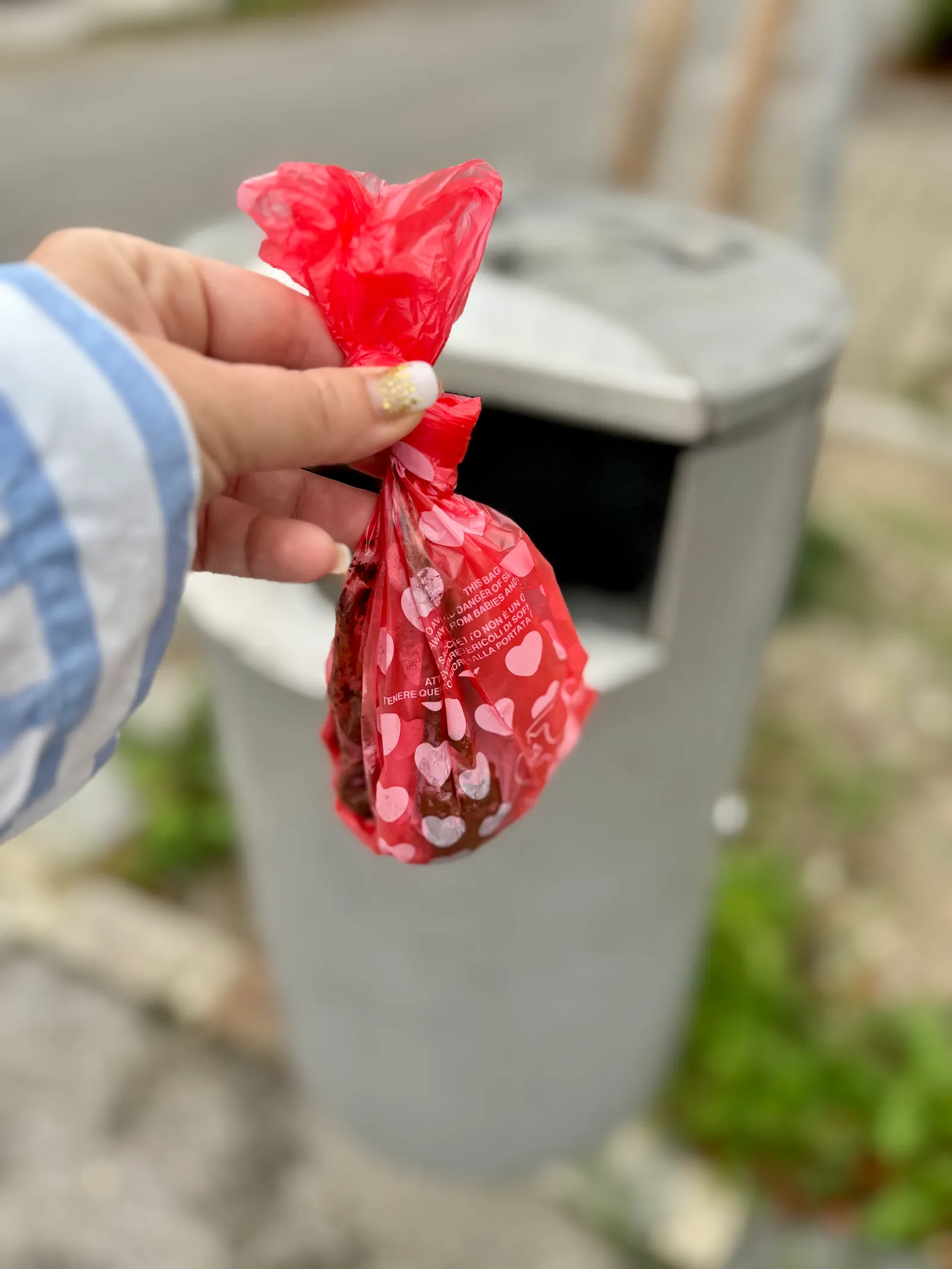 A hand holding a red dog waste bag in front of a grey rubbish bin on the street.