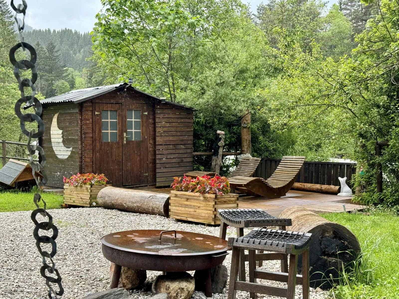 Gartenbereich mit Holzterrasse, Liegestühlen und Gartenhaus, im Vordergrund eine Feuerschale.