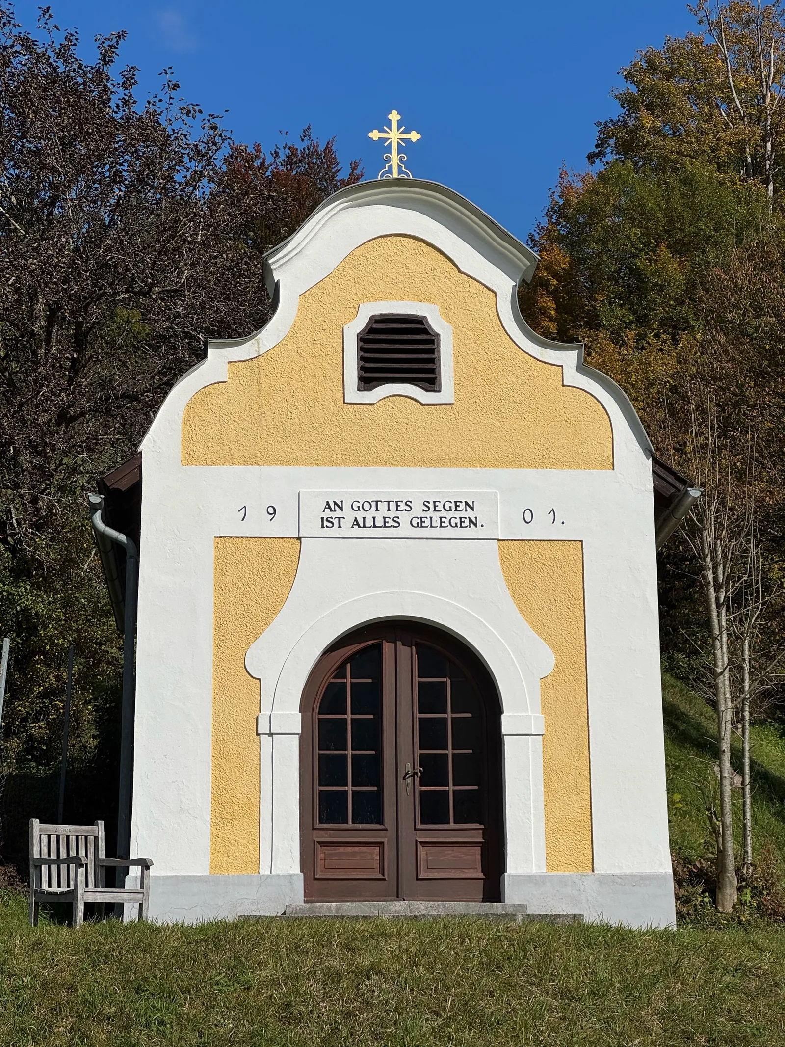 Kleine gelb-weiße Kapelle von 1901 mit Goldkreuz, umgeben von herbstlichen Bäumen.