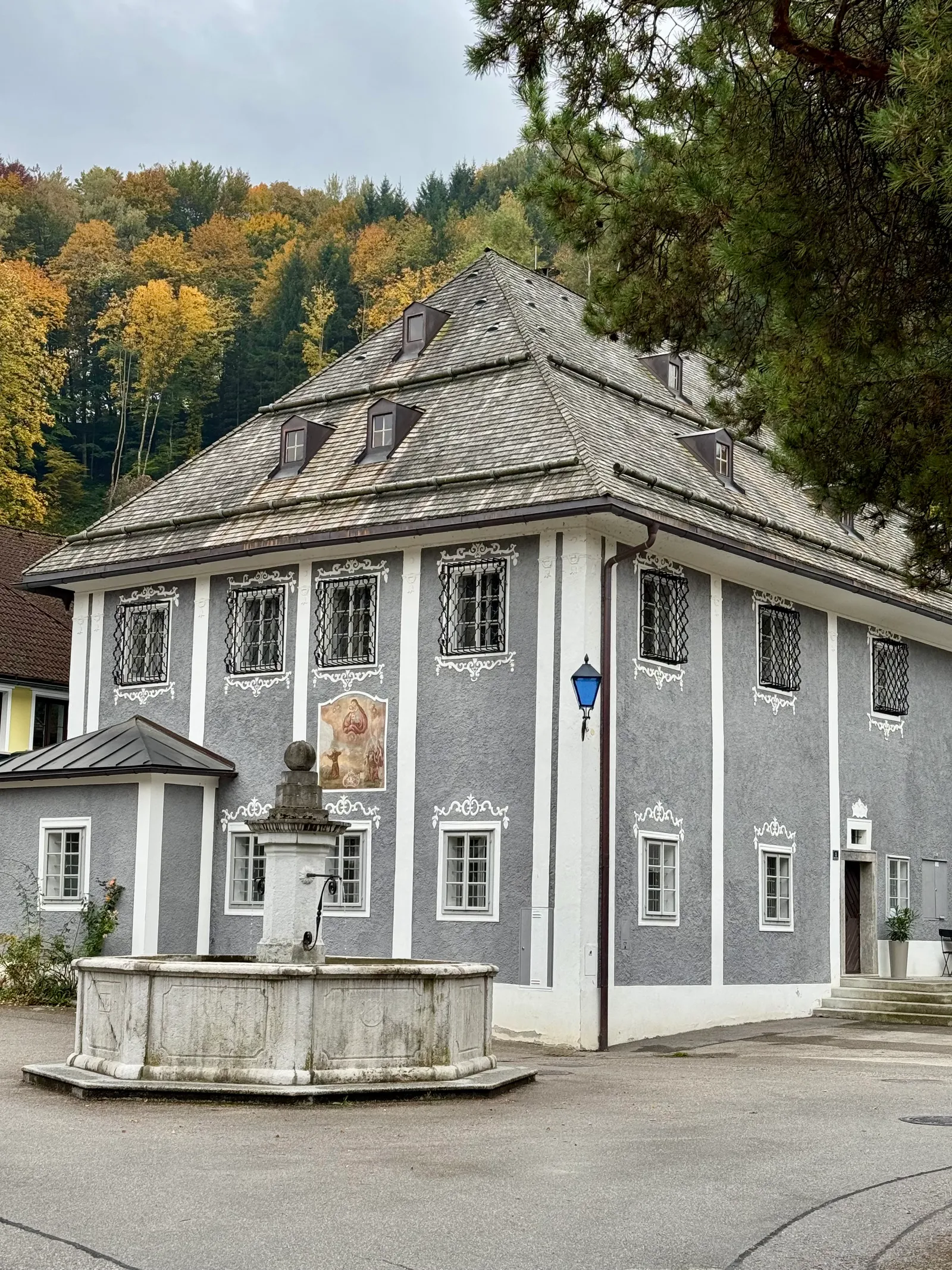 Barockes graues Gebäude mit weißen Stuckornamenten und Steinbrunnen davor in Ybbsitz, herbstliche Bäume im Hintergrund.