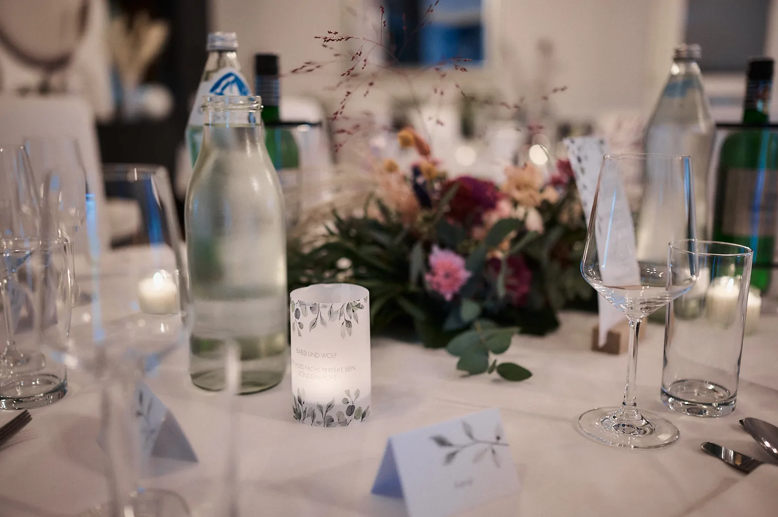 Hochzeitstisch mit personalisierten Kerzen, Blumengesteck, Gläsern und Wasserflaschen auf weißem Tischtuch.