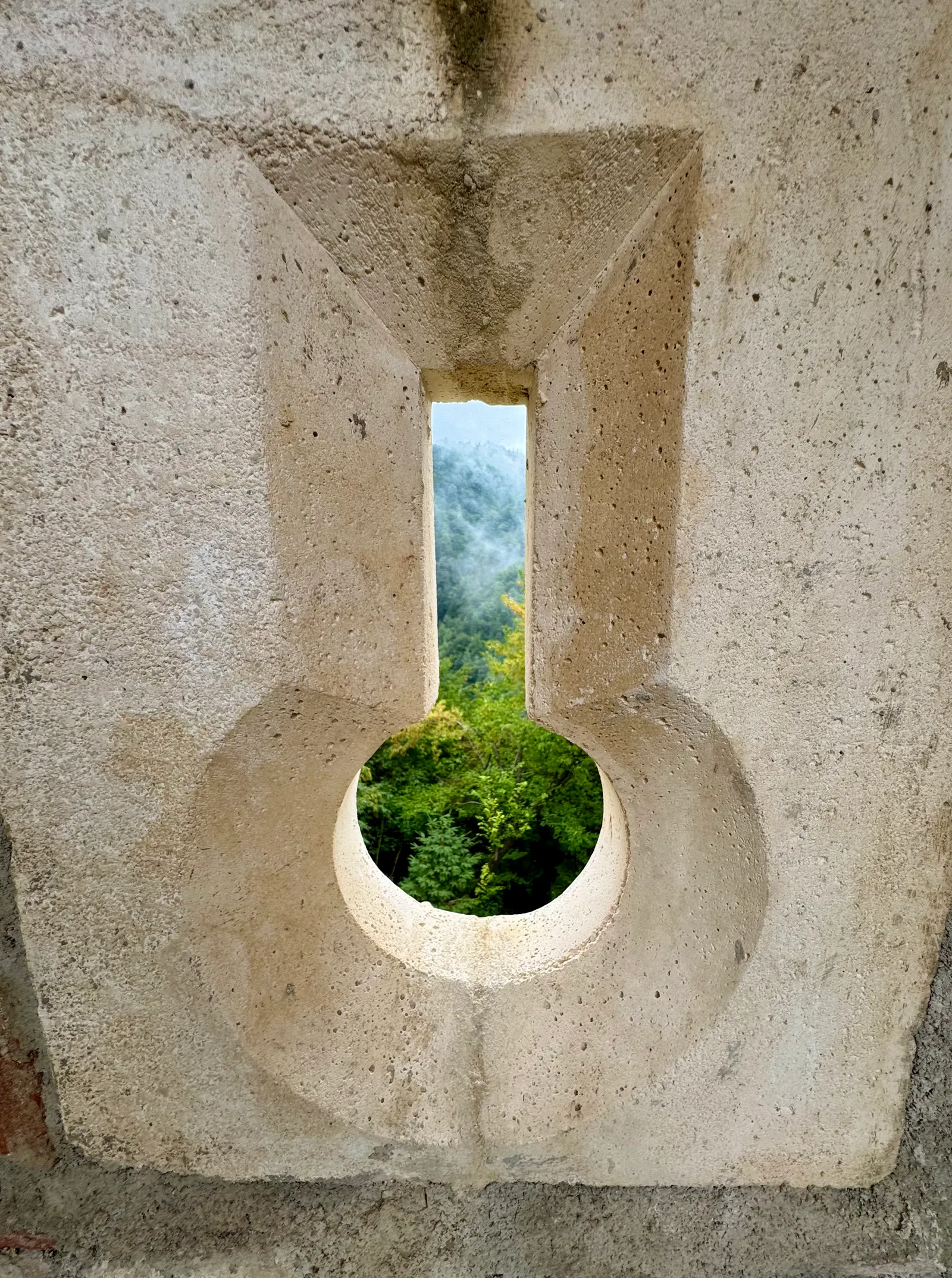 Schlüssellochförmige Schießscharte in der Burgruinenmauer mit Blick auf bewaldete Hügel im Nebel.