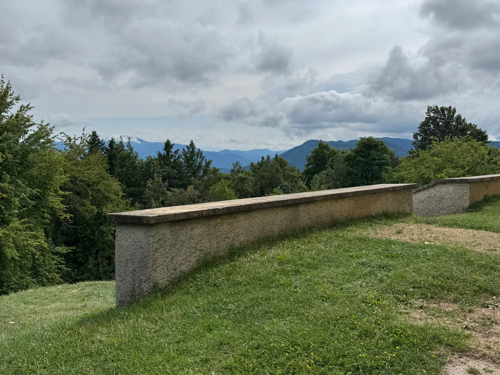 Steinbrüstung auf einer Anhöhe mit weitem Blick auf Berge und Wälder unter bewölktem Himmel.