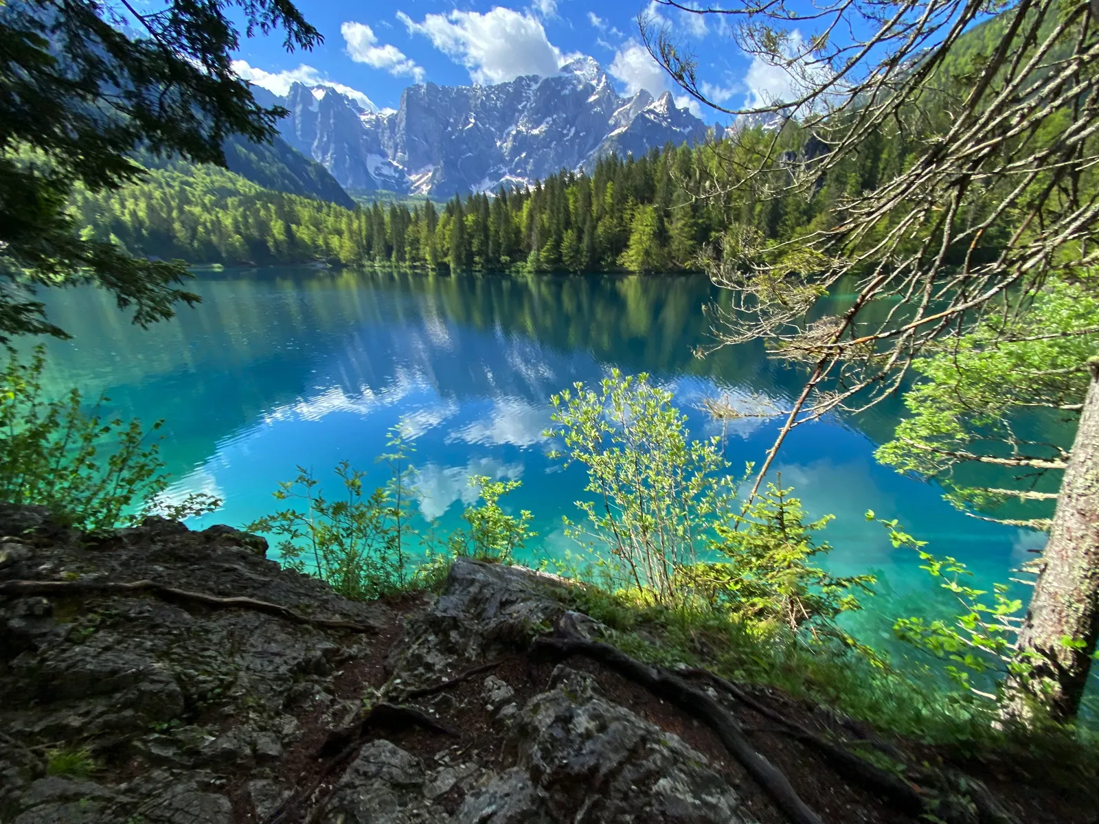 Türkisblauer Bergsee vom felsigen Ufer aus, Wald und Berge spiegeln sich im Wasser.