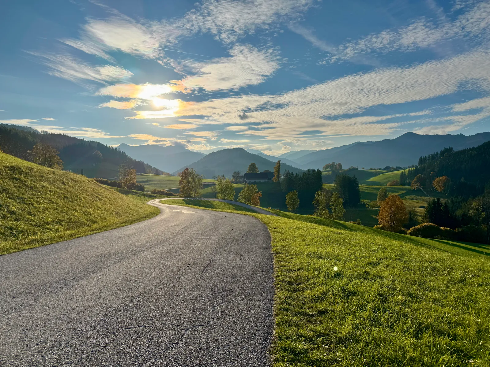 Kurvenreiche Landstraße durch hügelige Herbstlandschaft mit Bergen und tief stehender Sonne.