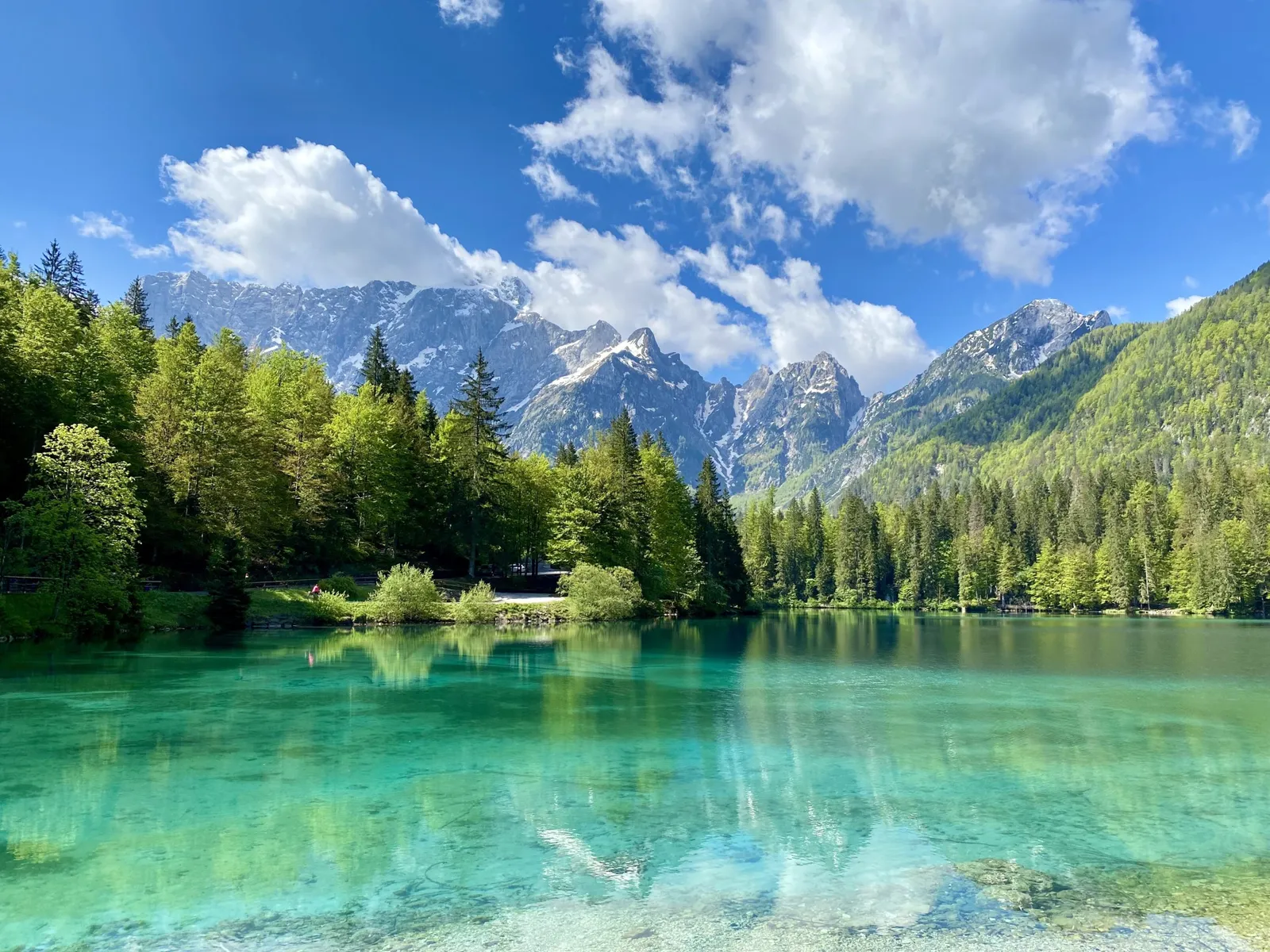 Türkisfarbener Bergsee mit Nadelwald und schneebedeckten Gipfeln im Hintergrund.