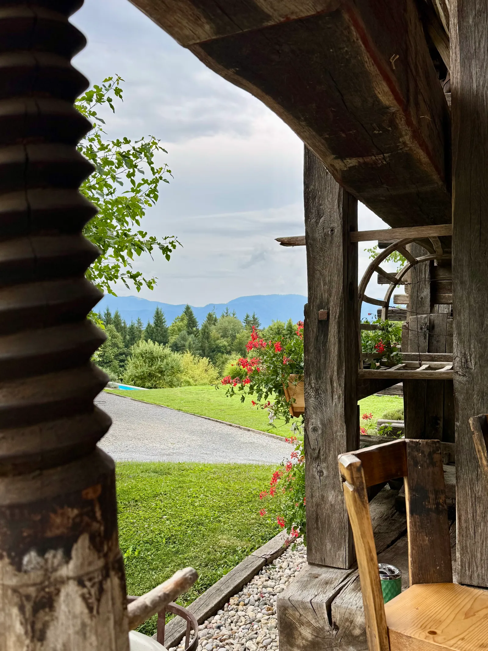Blick durch altes Holzgebälk auf Rasen, rote Blüten und Berge im Hintergrund.