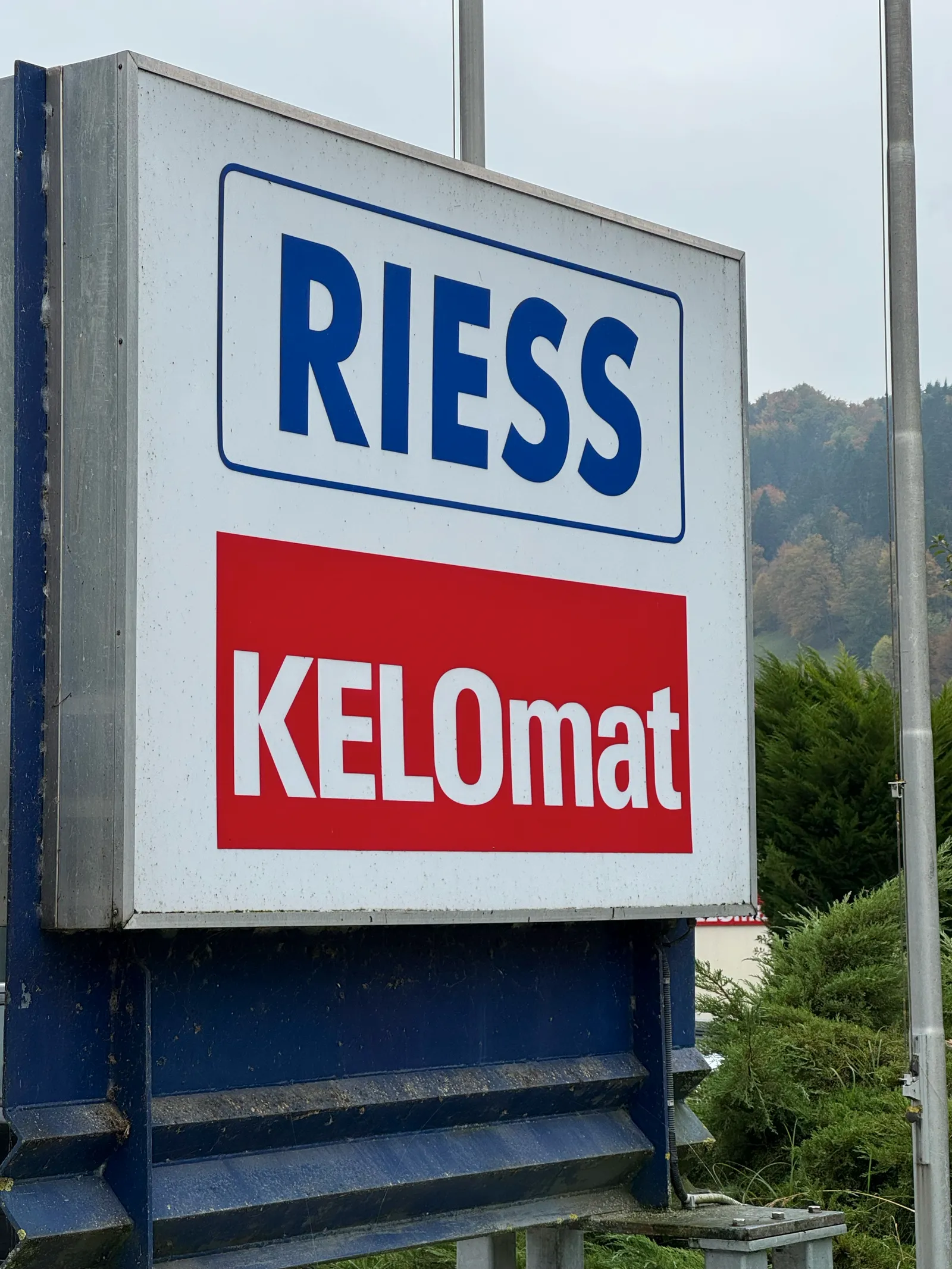 Firmenschild von Riess und KELOmat auf einer blauen Stele, herbstlicher Wald dahinter.