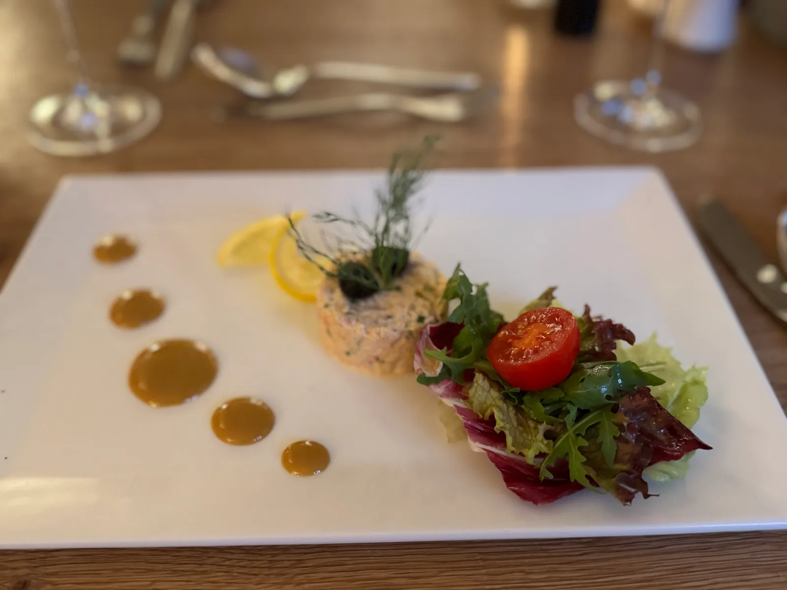 Lachstatar mit Blattsalat und Senfsauce-Tupfen auf einem weißen Teller.