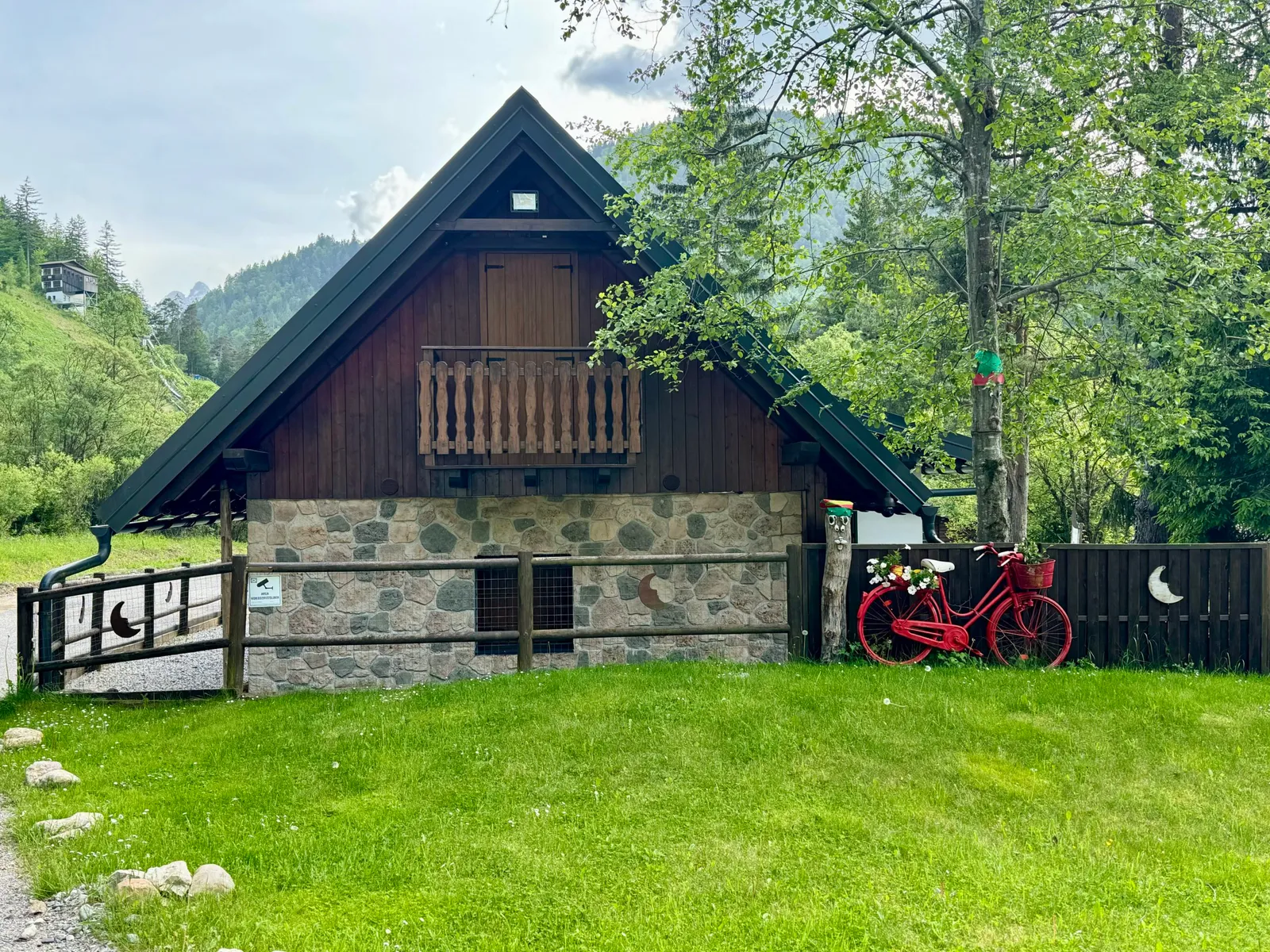 Holzchalet mit Steinsockel und rotem Fahrrad als Dekoration an einem Zaun.