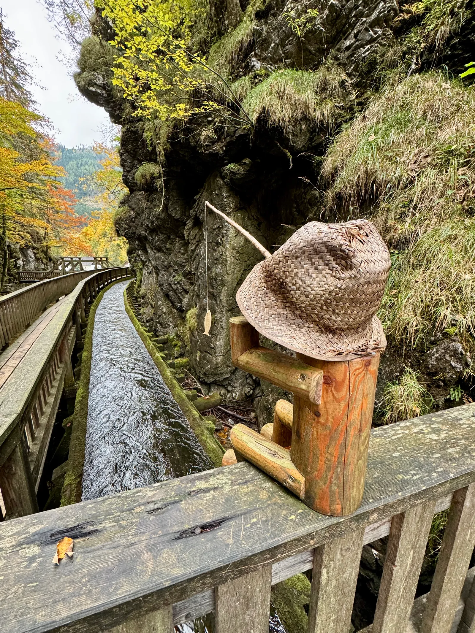 Strohhut und kleines Holzmännchen mit Angelrute auf dem Geländer eines Stegs über dem Bach in der Schlucht.