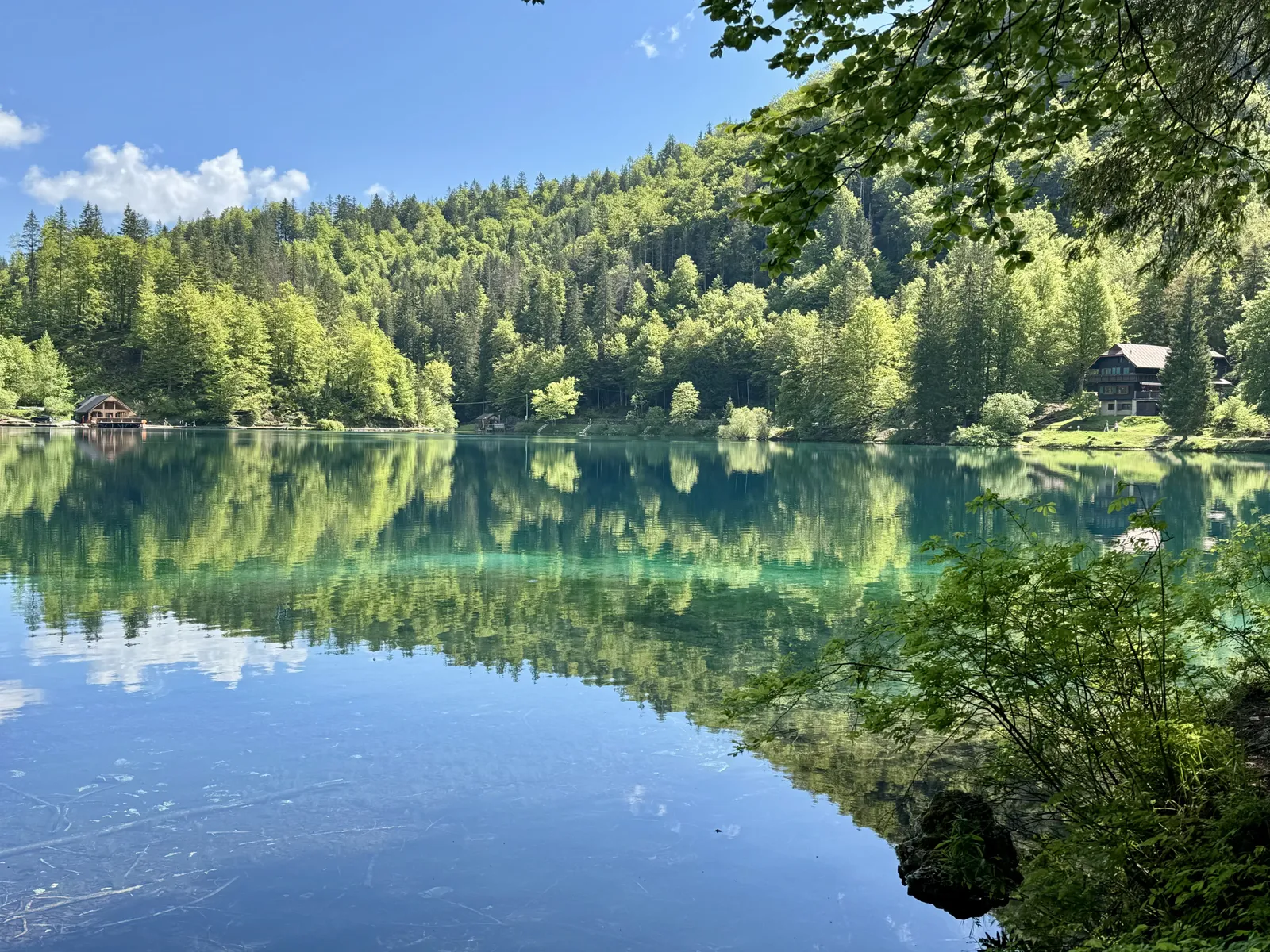 Ruhiger Bergsee mit Waldspiegelung und Holzhäusern am gegenüberliegenden Ufer.