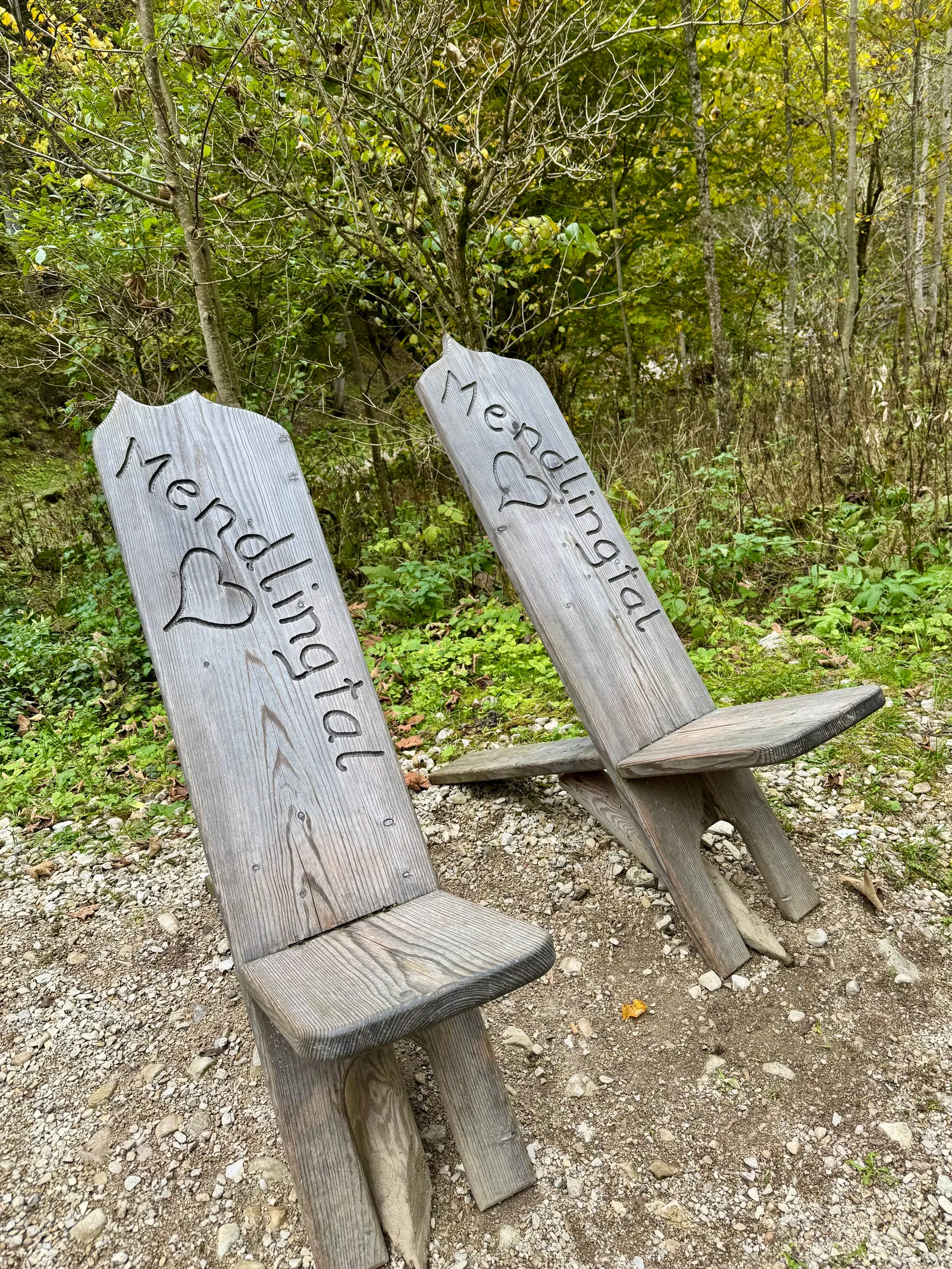 Zwei geschnitzte Holzsessel mit der Aufschrift „Mendlingtal" stehen auf Kies vor herbstlichem Wald.