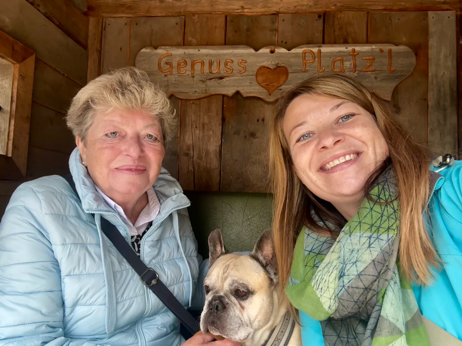 Babsi, ihre Mutter und Murphy sitzen unter einem Holzschild mit der Aufschrift „Genuss Platzl".