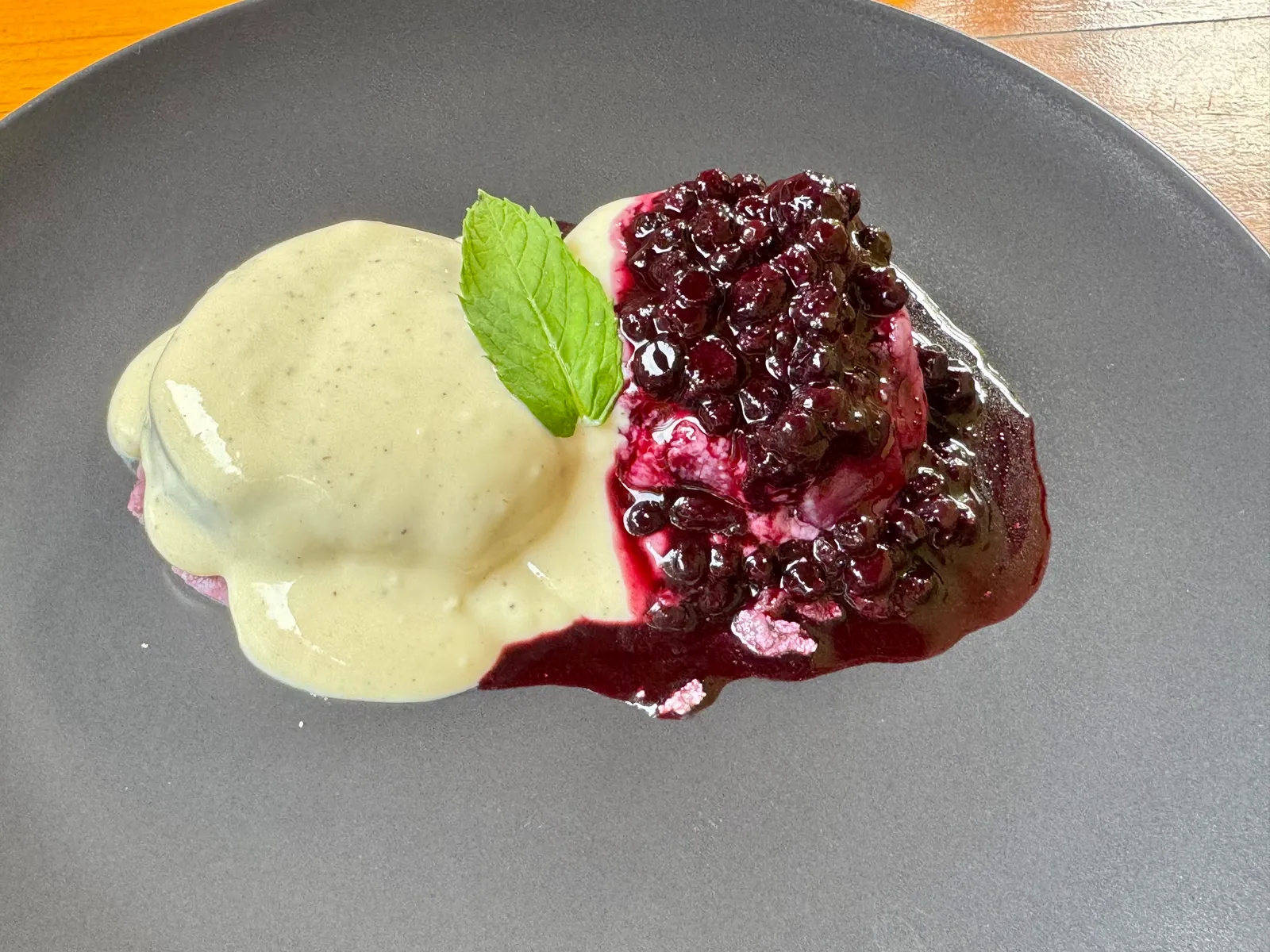 Dessert mit Vanillesauce und dunklem Beerenkompott auf einem grauen Teller, Minze als Garnitur.