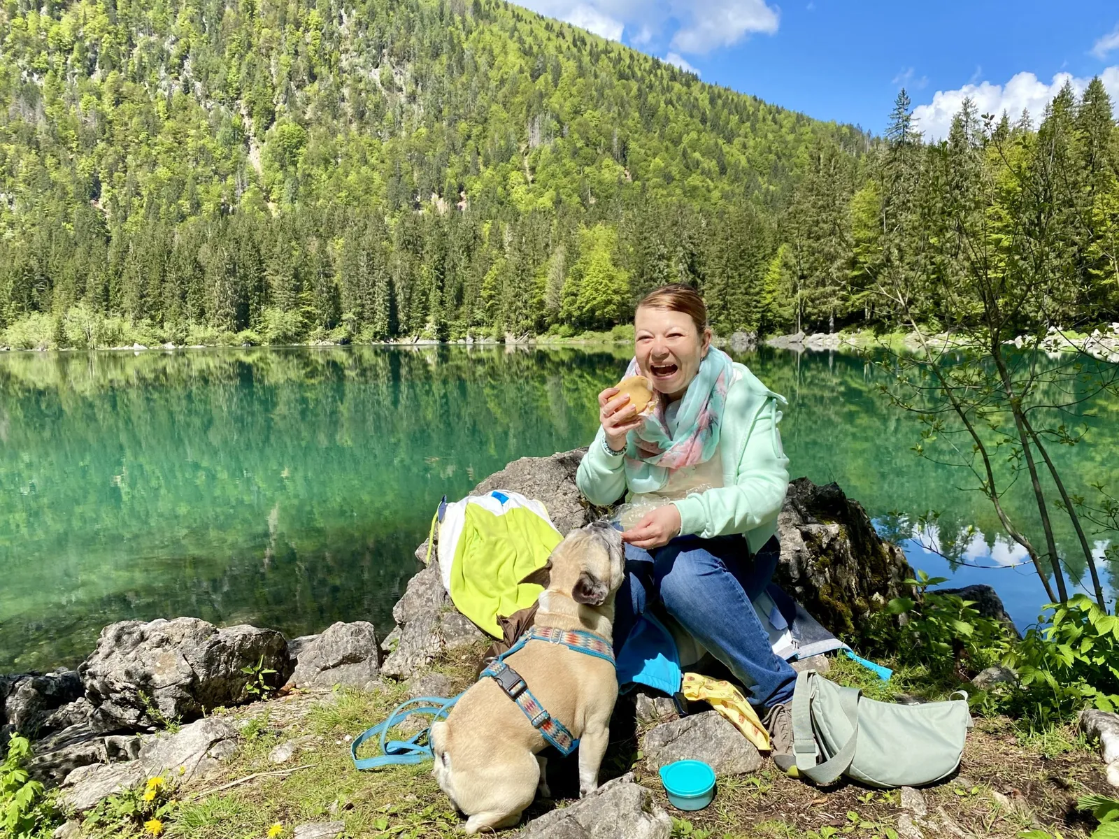 Eine Frau isst am Seeufer auf einem Felsen, eine Französische Bulldogge sitzt daneben.