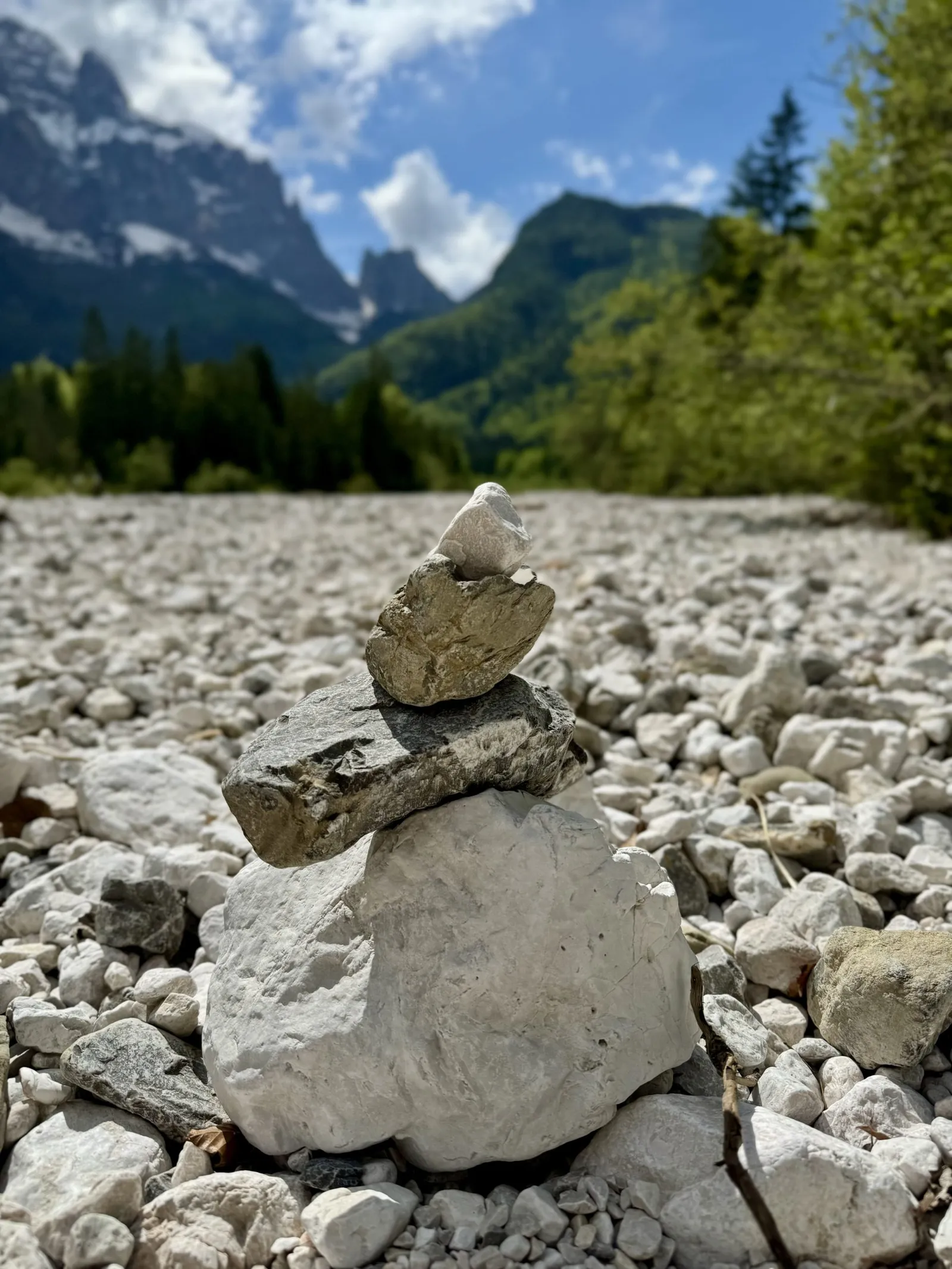 Eine Steinpyramide auf einem Schotterbett im Val Saisera, dahinter Bergwände und Wald.