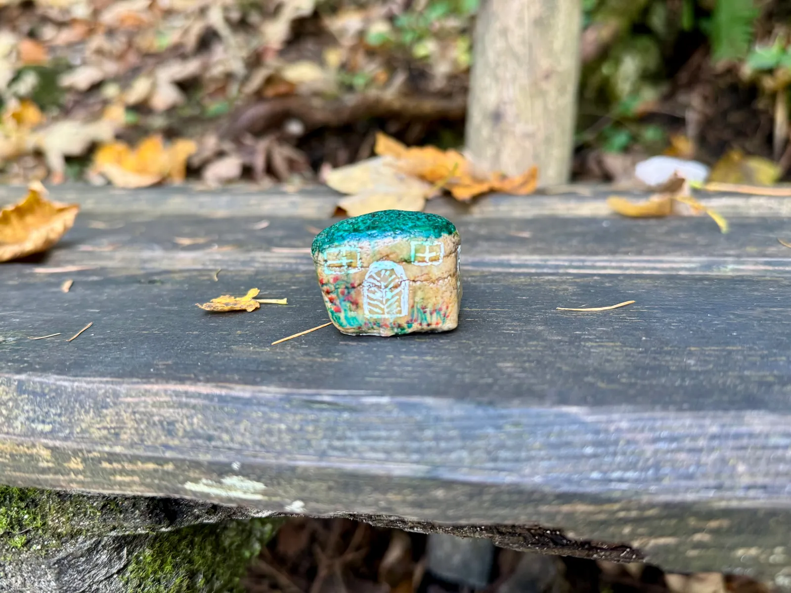Ein bemalter Stein in Hausform liegt auf einer Holzbank im herbstlichen Wald.