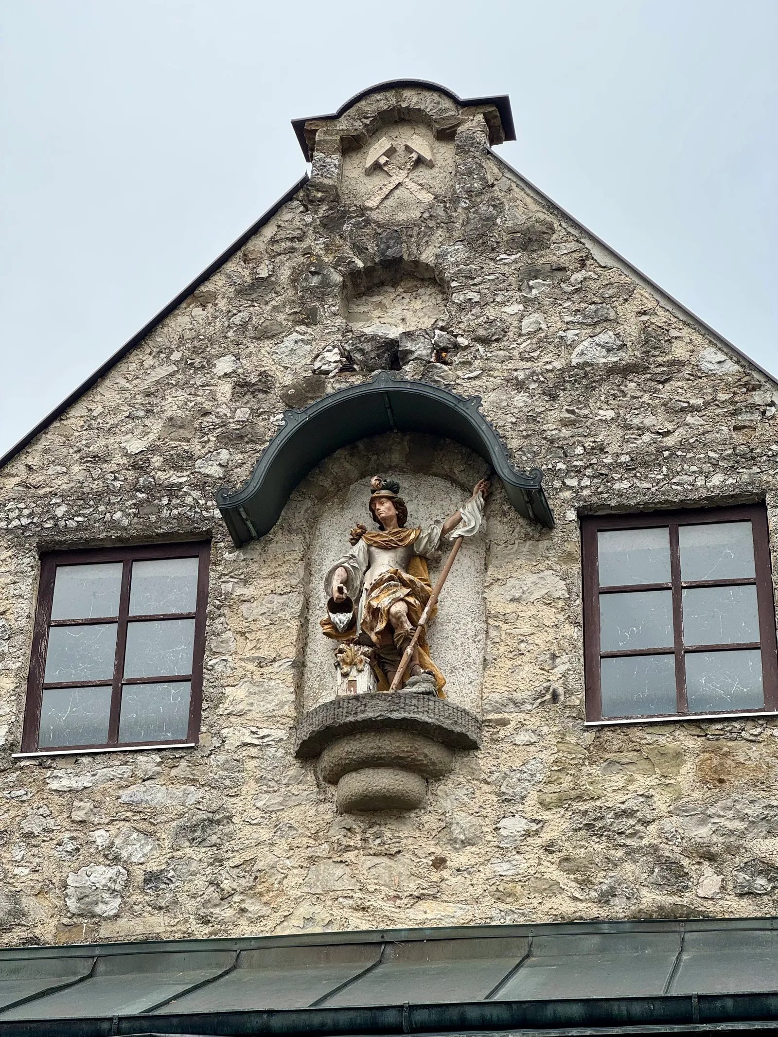 Bemalte barocke Heiligenfigur in einer Wandnische eines Steingebäudes, darüber ein Bergmann-Symbol.