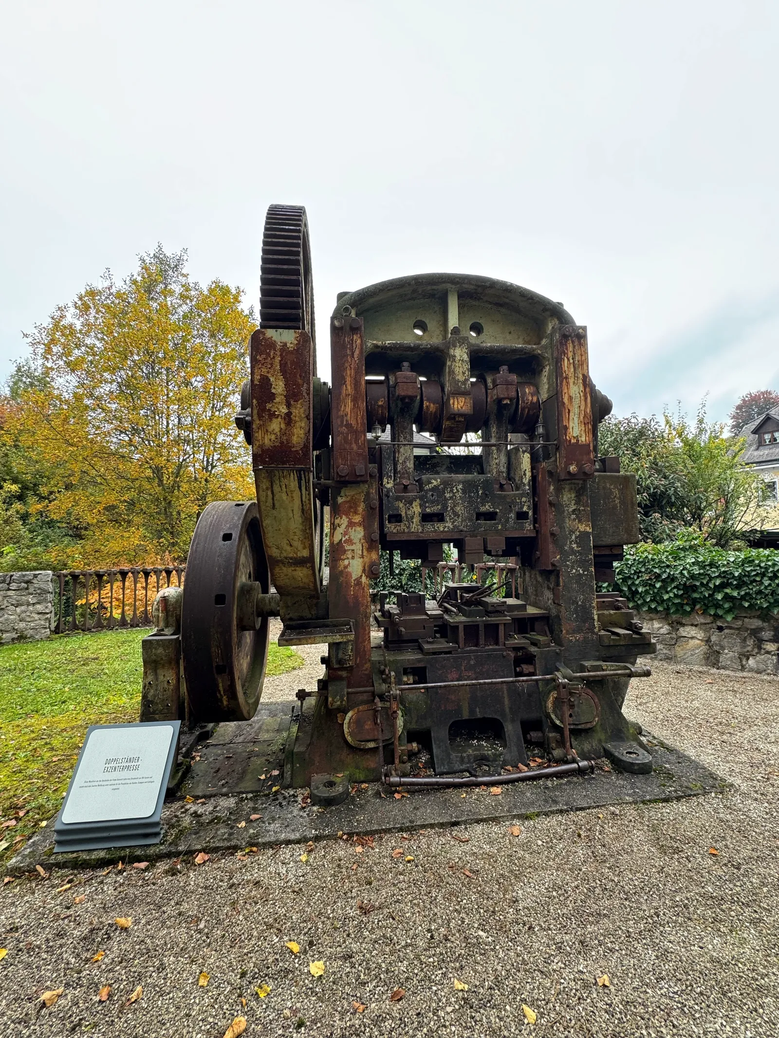Historische Exzenterpresse aus Eisen als Freiluftexponat in einem Garten, herbstliche Bäume dahinter.