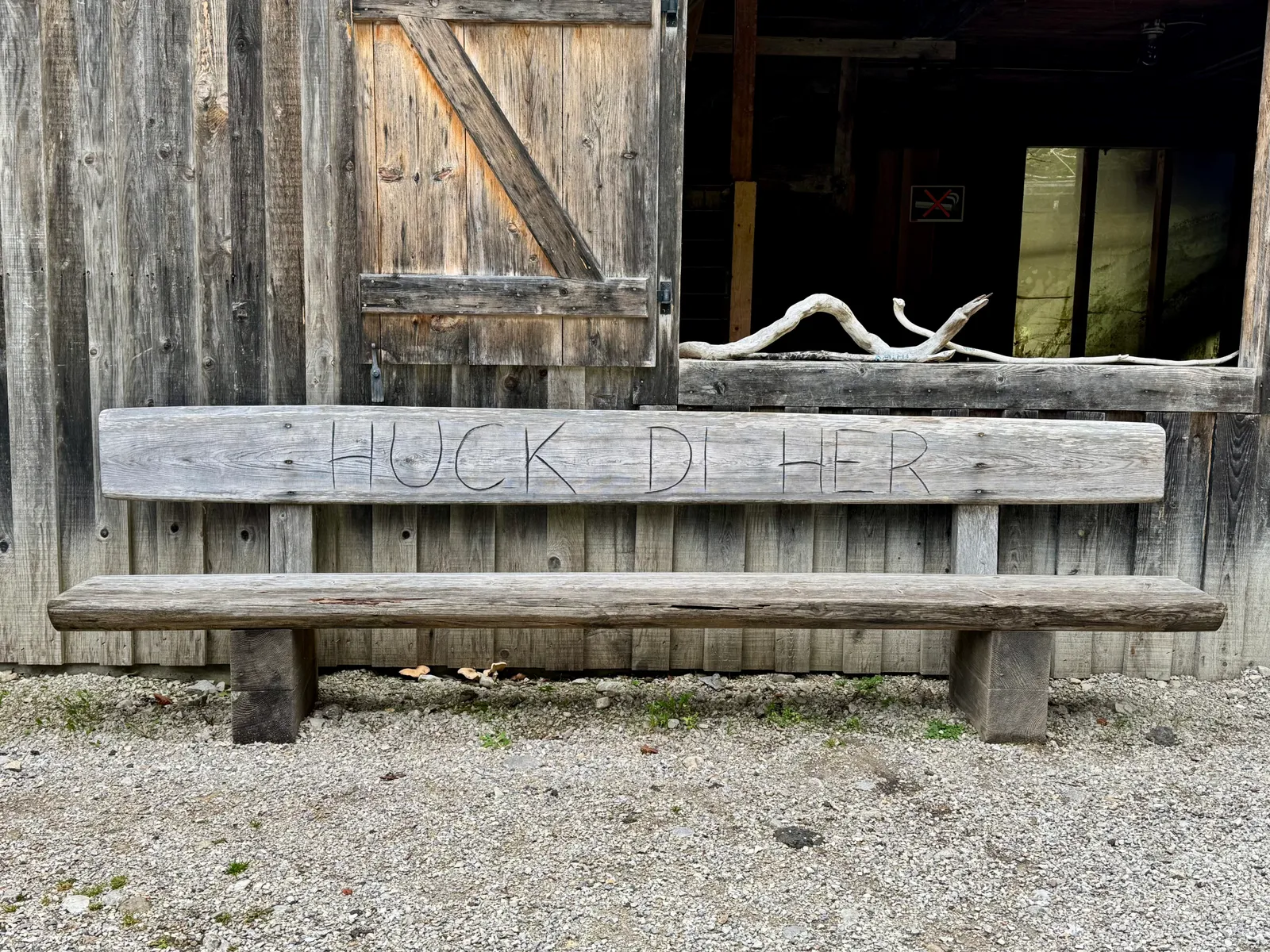 Holzbank mit eingeschnitztem Spruch „Huck di her" vor einer alten Scheunentür aus verwittertem Holz.