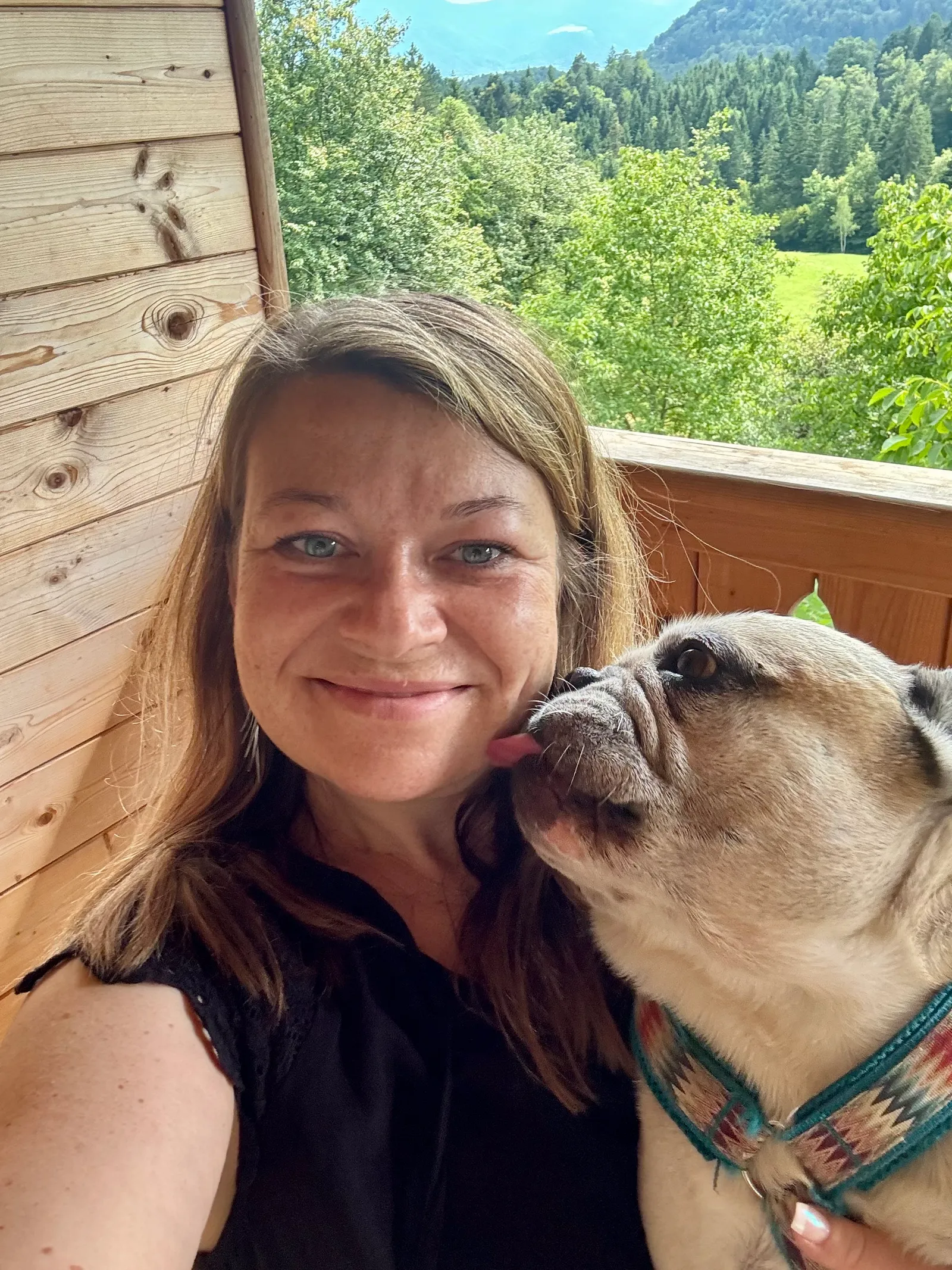 Murphy leckt Babsi auf einem Holzbalkon die Wange, grüner Wald im Hintergrund.